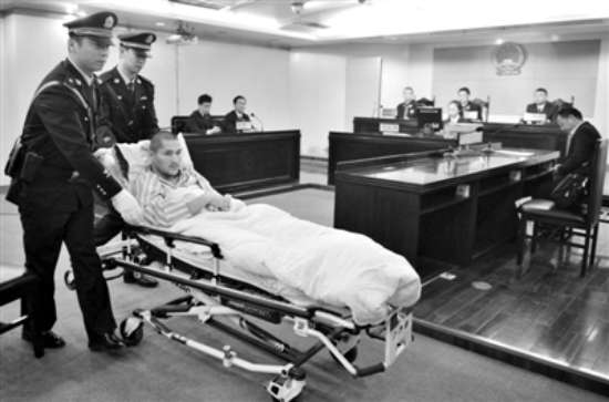 图为：2013年10月15日，“首都机场7·20爆炸案”在北京市朝阳区人民法院一审宣判，冀中星剃了光头，躺在病床上，由两名民警推着进法庭现场。