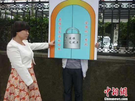 DAY12.jpg 图为:北京西单大木仓胡同的教育部大院门前,只见志愿者高举一块代表大门的塑料板,门楣上标有“普通高等学校招生全国统一考试”的字样。河南新乡籍视障人士王瑞从一旁走向这扇“高考之门”,却因大门紧锁而碰壁。门锁上挂有“不提供盲文试卷”的告知牌。